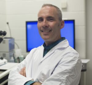 John Peever is wearing a lab coat and is standing in front of a computer monitor and other equipment in a lab.